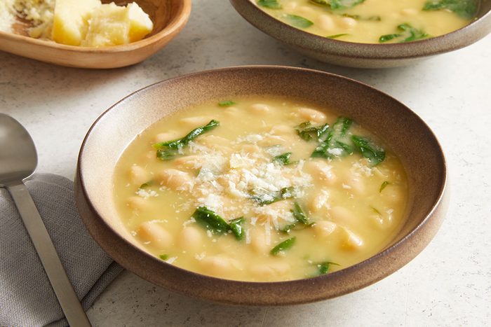 A bowl of creamy white bean soup with spinach, topped with grated cheese, sits on a table next to a spoon and a plate of sliced bread with butter.