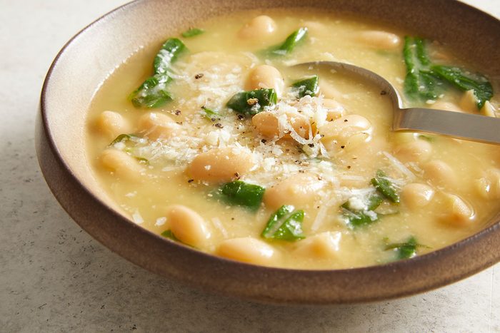A bowl of creamy white bean soup with spinach, topped with grated cheese and black pepper, with a spoon resting in the soup.