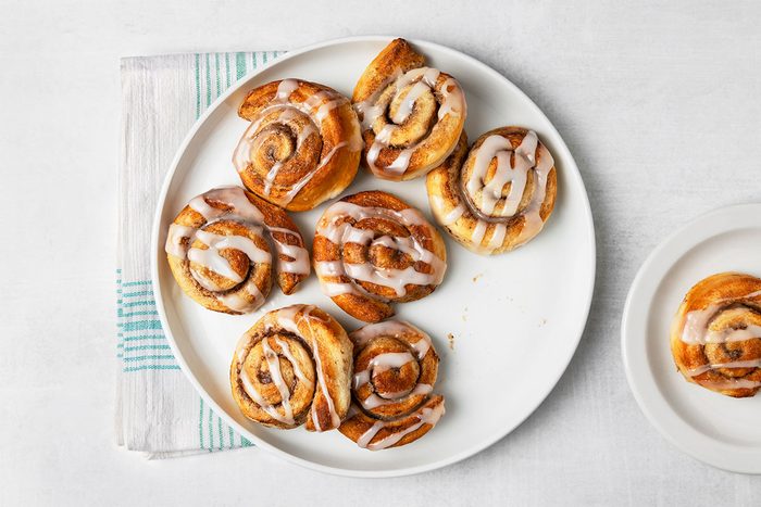 A white plate with seven icing-drizzled cinnamon rolls on a white surface, next to a striped cloth and a smaller plate holding one additional cinnamon roll.