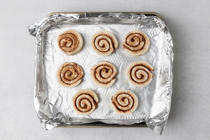 Eight unbaked cinnamon rolls are arranged in three rows on a baking tray lined with crumpled aluminum foil. The cinnamon rolls are evenly spaced on the foil surface.