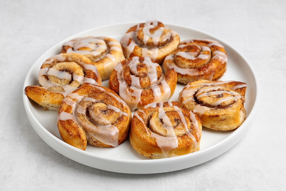 A white plate with eight cinnamon rolls topped with white icing, arranged neatly on a light-colored surface.