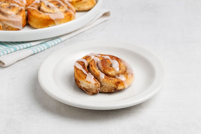 Single iced cinnamon roll sits on a white plate, with more cinnamon rolls on a nearby dish in the background. The setting is simple and light-colored. 279480 Christinema 7
