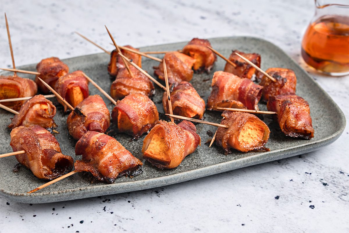 A gray rectangular plate holds several bacon-wrapped sweet potato bites secured with toothpicks. A small glass container of syrup sits in the background on a light, speckled surface.