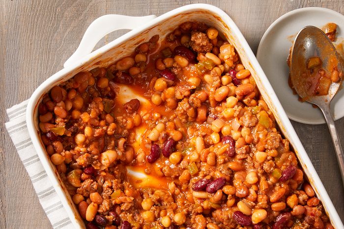 A white baking dish filled with baked beans and ground meat in tomato sauce, partially served on a plate beside it with a serving spoon. The dish rests on a striped cloth on a wooden surface.