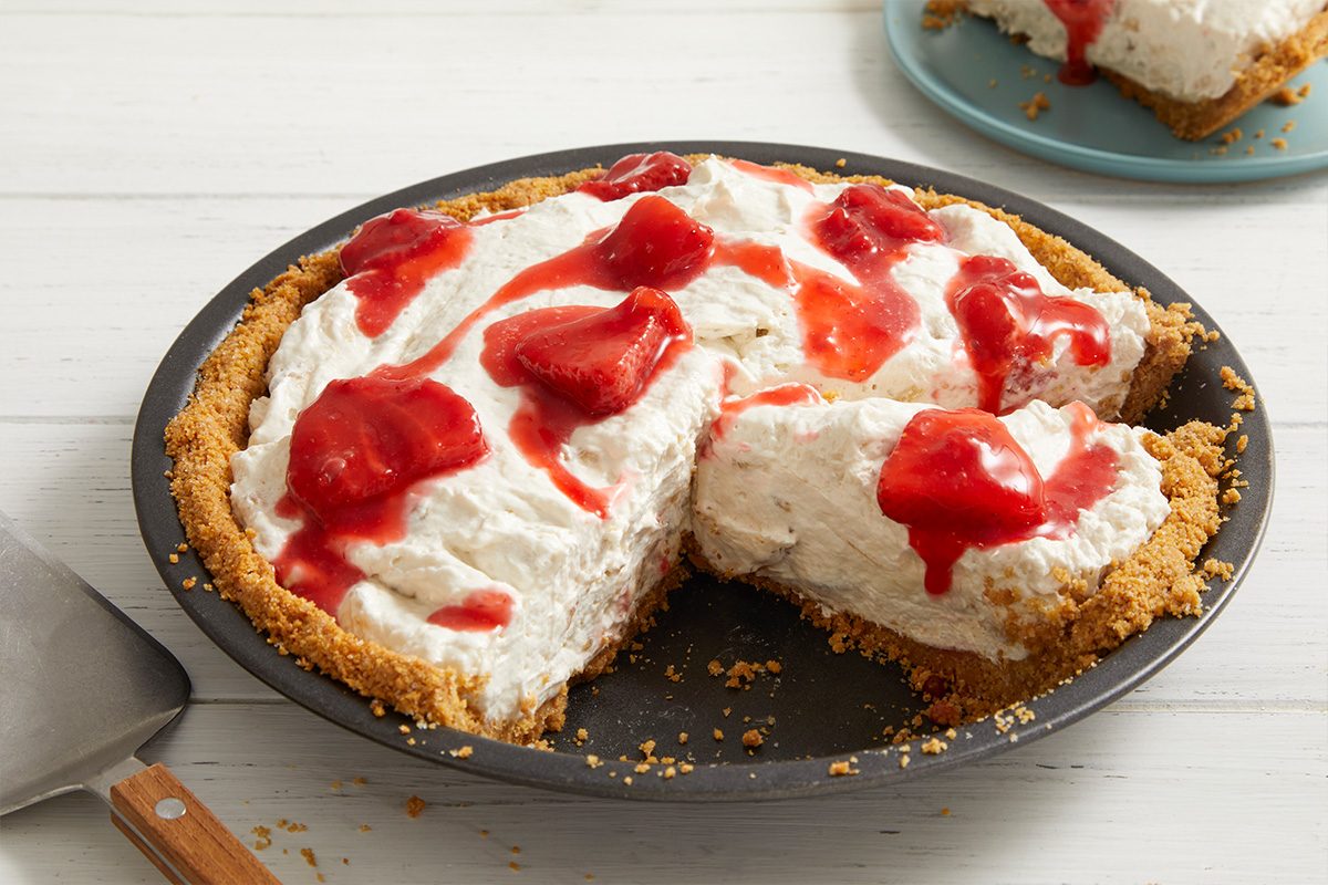 A creamy pie with a graham cracker crust, topped with whipped filling and strawberry sauce, sits on a white wooden table. One slice is missing, and the slice is served on a blue plate in the background.