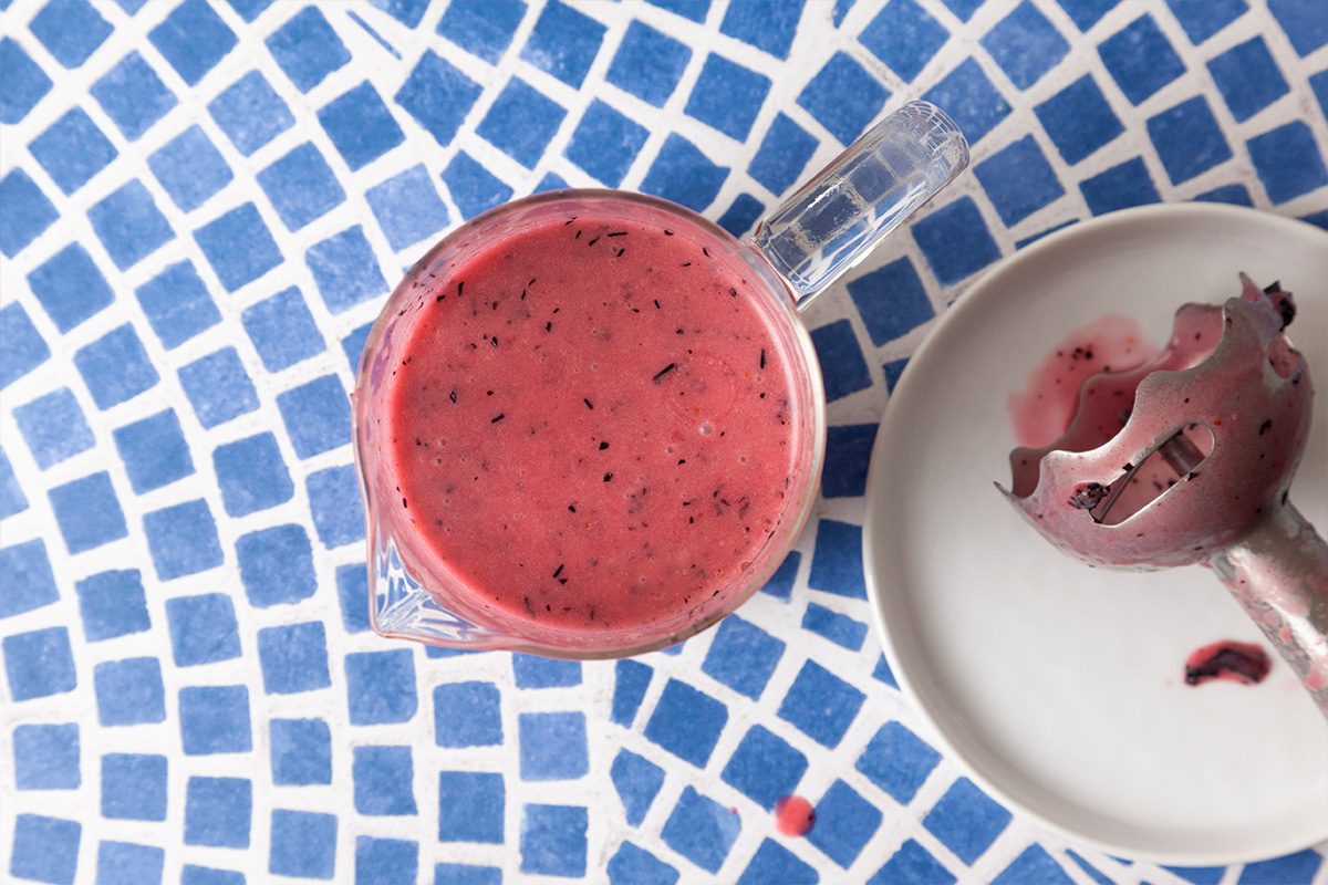 A glass cup filled with a thick, pink berry smoothie sits on a blue and white mosaic table next to a plate holding a partially used ice cream scoop stained with smoothie.