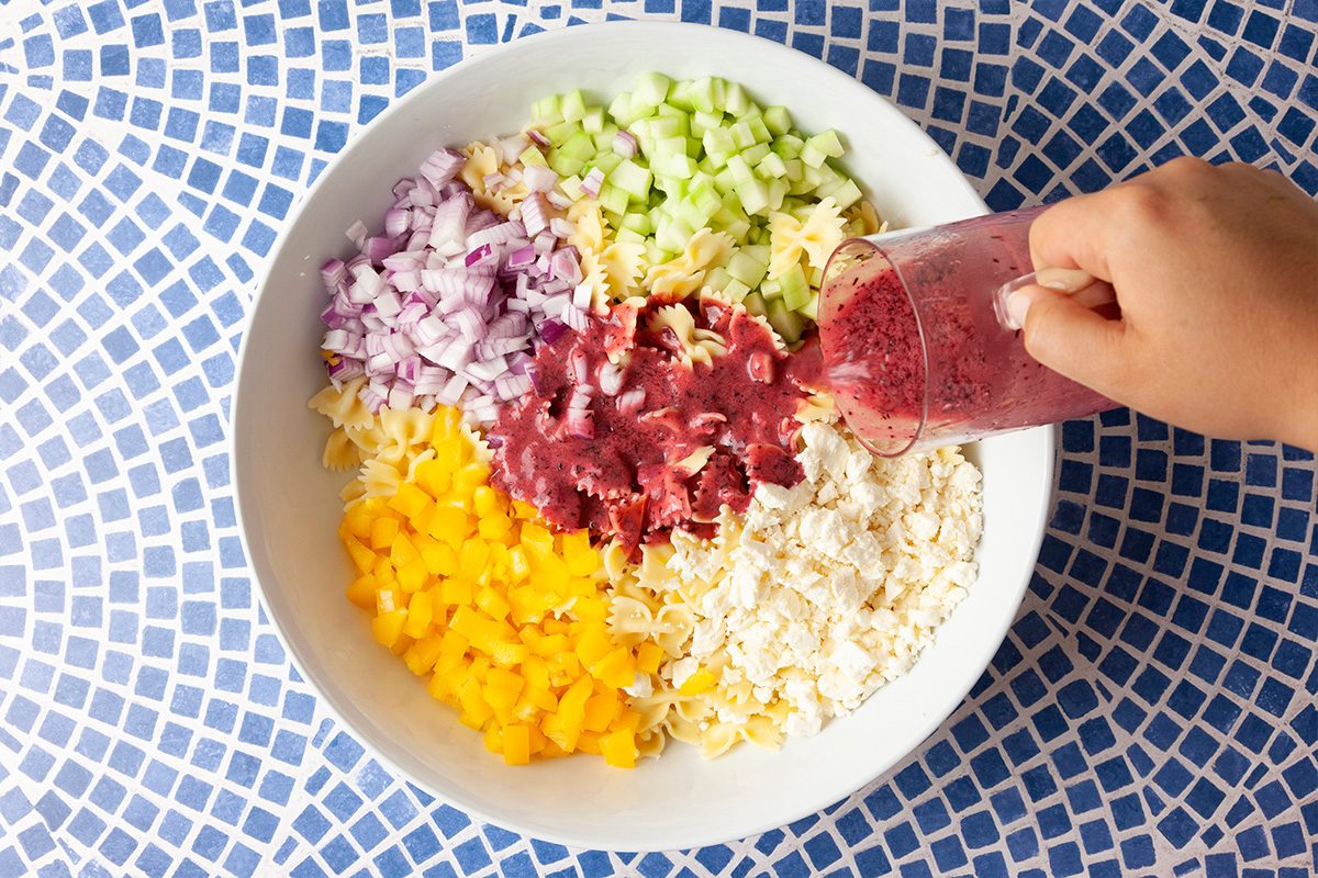 A hand pours a red-purple dressing over a bowl filled with chopped red onion, celery, yellow bell pepper, rice, and pineapple, all arranged in sections on a blue and white mosaic tile table.