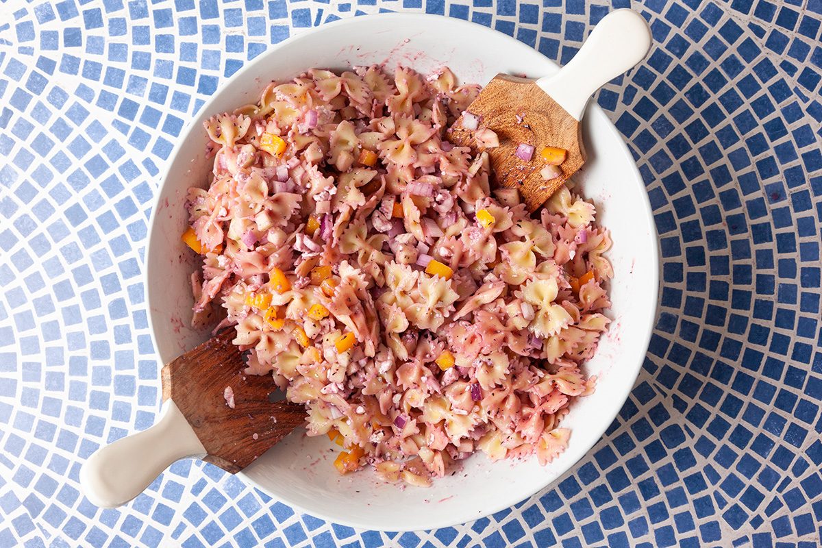 A bowl of colorful pasta salad with bowtie pasta, chopped red onion, and orange bell pepper, mixed together and served with wooden utensils on a blue and white mosaic-patterned table.