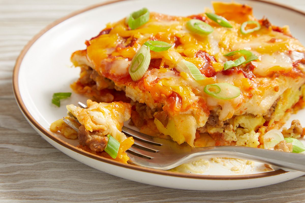 A close-up of a plate with a slice of cheesy lasagna topped with chopped green onions. A fork rests on the plate, with a bite of lasagna on it. The background is a light, wooden table.