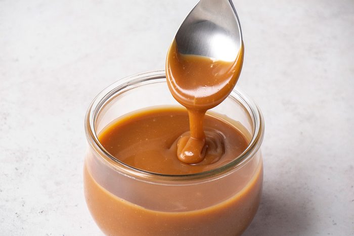 A spoon drizzles thick, golden caramel sauce into a small glass jar filled with caramel, set on a light-colored surface.