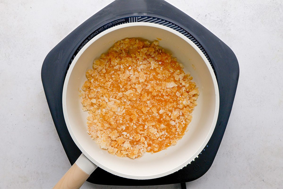 A saucepan with light brown, crumbly melted sugar sits on a black hot plate against a light background. The sugar is partially liquefied and has a grainy texture.