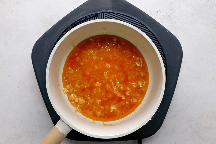 A white saucepan with a wooden handle sits on a black hot plate, filled with a chunky orange mixture, possibly cooking fruit or sauce, against a light gray background.