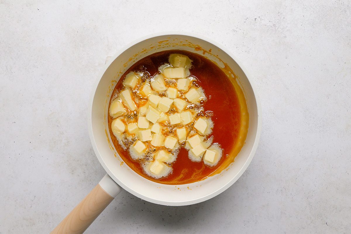 A white frying pan with a wooden handle contains melted caramel and cubes of butter, sitting on a light-colored surface.