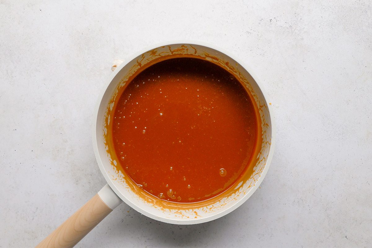 A white saucepan with a light wooden handle contains smooth, reddish-brown sauce. The pan sits on a light, textured surface.