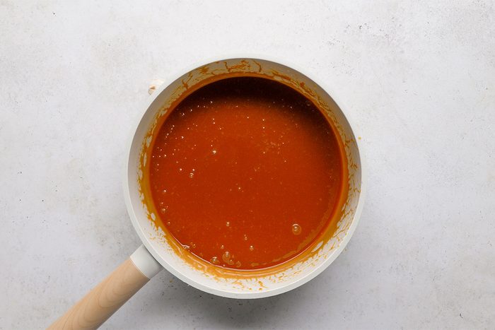 A white saucepan with a light wooden handle contains smooth, reddish-brown sauce. The pan sits on a light, textured surface.
