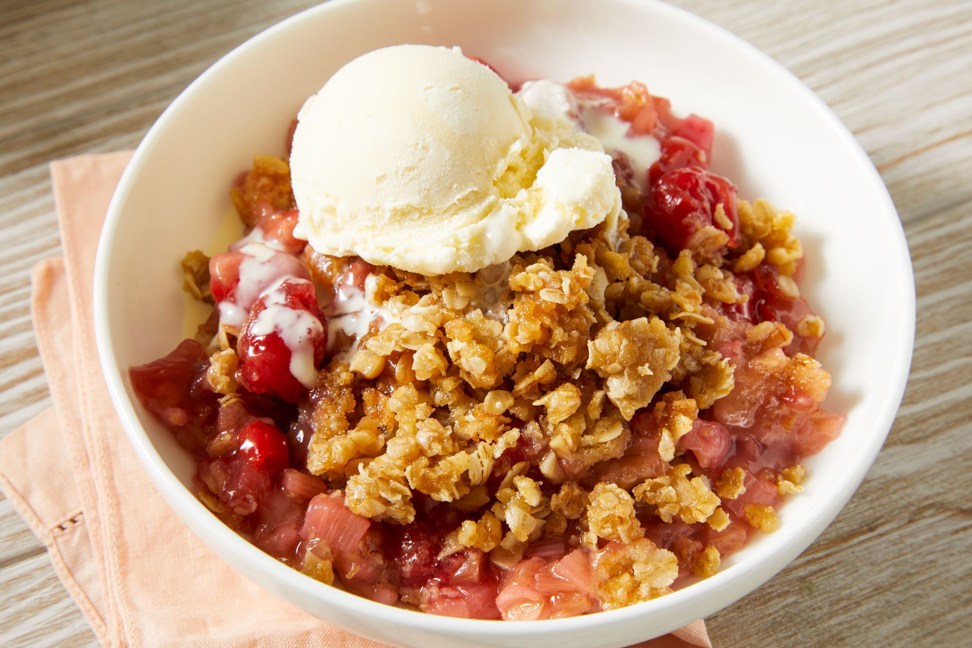 Close-up shot of Cherry Rhubarb Crunch; served in a bowl with ice cream; placed over a napkin; set on a cream-colored wooden surface