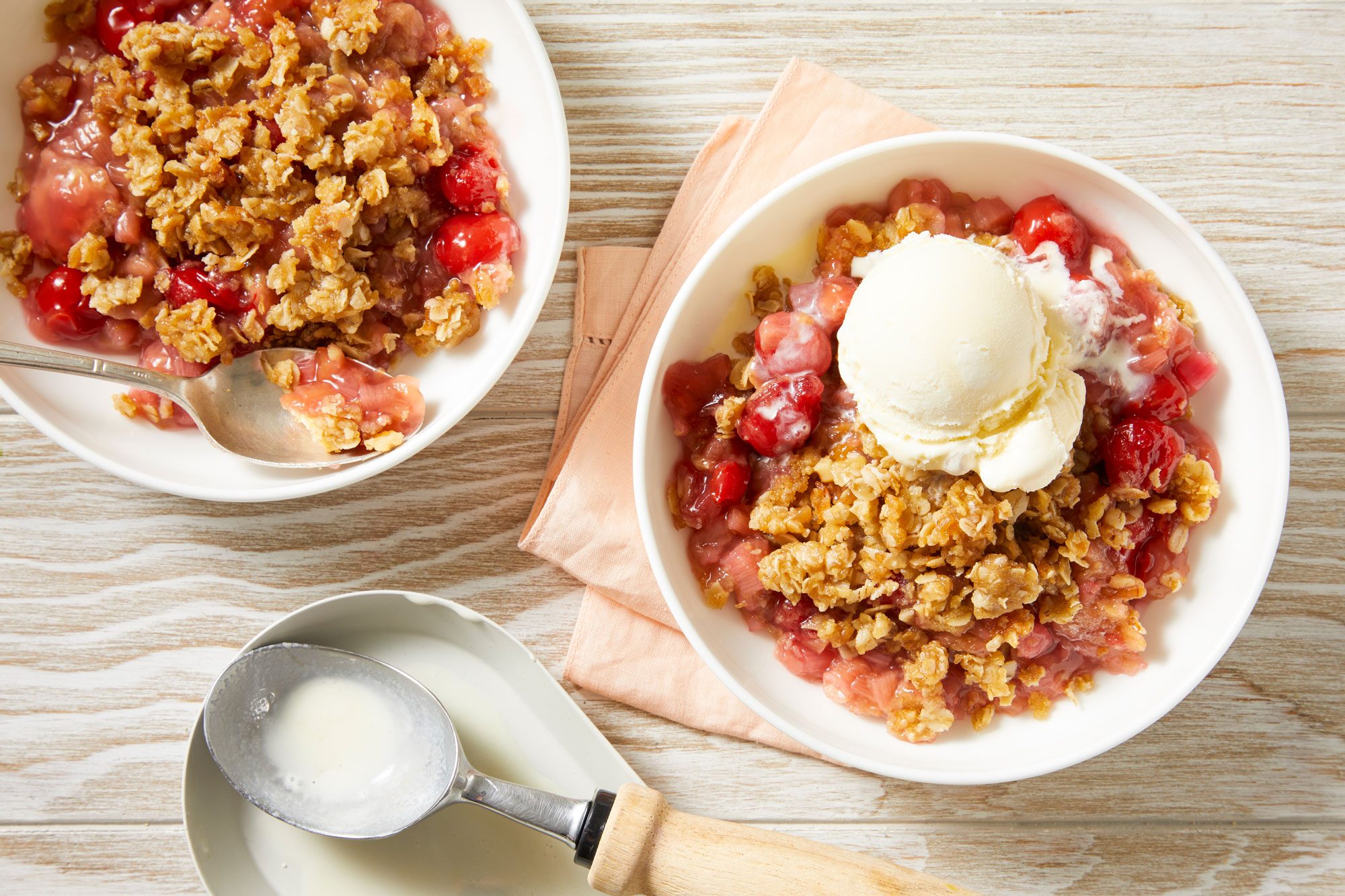 Overhead shot of Cherry Rhubarb Crunch; served in two bowls with ice cream; one bowl placed on a napkin, the other directly on a cream-colored wooden surface