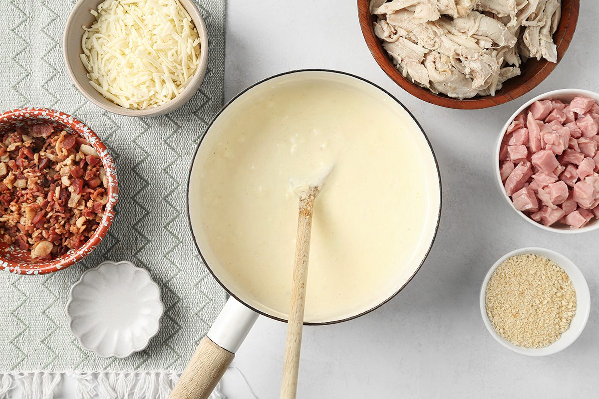 A saucepan of creamy white sauce with a wooden spoon is surrounded by bowls of shredded cheese, cooked chicken, diced ham, chopped bacon, and breadcrumbs on a light surface.