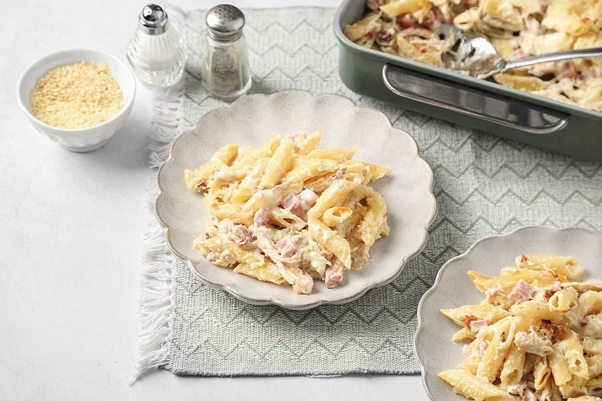 A plate of creamy pasta with ham is served on a white scalloped dish, with more pasta in a baking dish nearby; a small bowl of grated cheese and salt and pepper shakers are also on the textured cloth.
