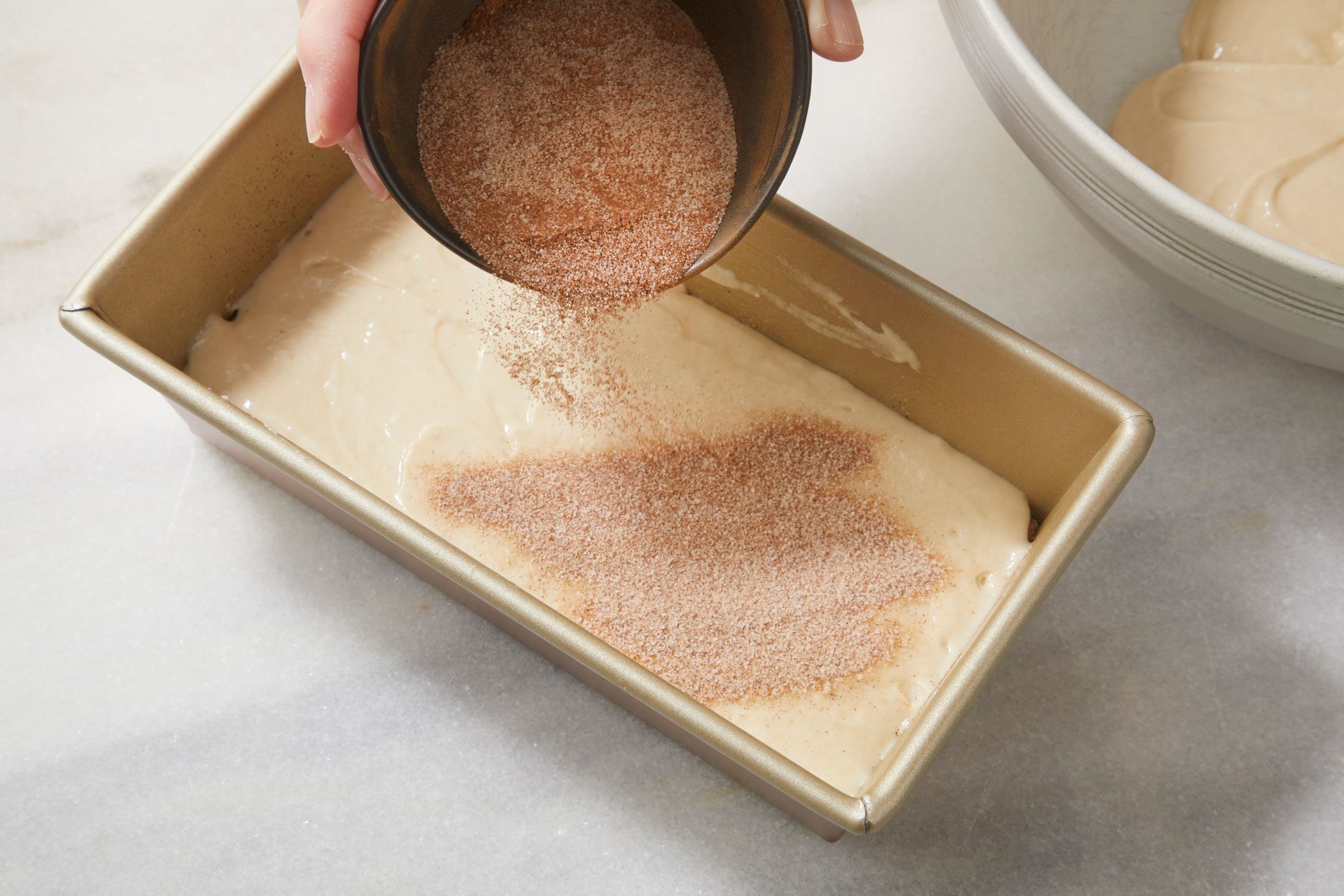 overhead shot of a hand sprinkles cinnamon sugar from a small bowl onto unbaked batter in a rectangular loaf pan, with a mixing bowl in the background