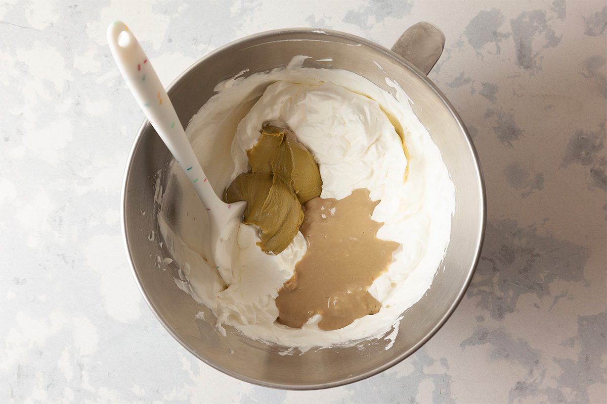 A metal mixing bowl containing white creamy mixture, pickle slices, and a light brown liquid, with a white spatula resting inside the bowl on a light-colored surface.