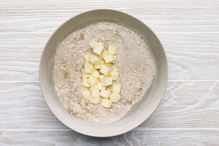 A mixing bowl containing flour mixture and several small cubes of butter, set on a light wooden surface.