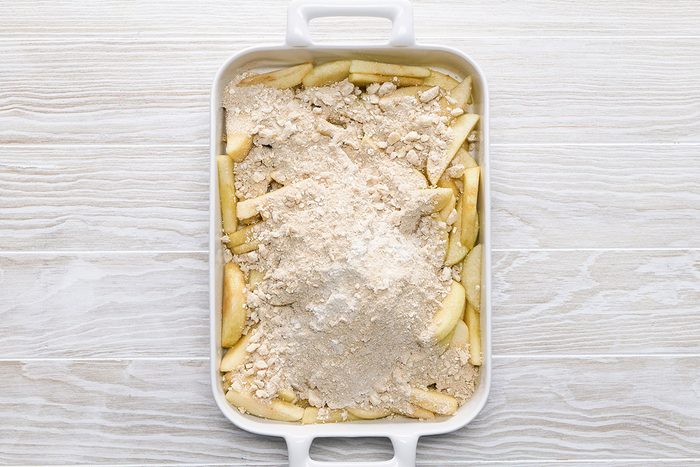 A white baking dish filled with sliced apples topped with an even layer of dry crumble mixture, ready to be baked, sits on a light wood surface.
