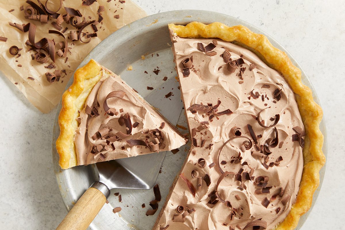 A chocolate cream pie in a metal dish, topped with chocolate shavings. One slice is cut and being lifted with a pie server onto a plate nearby. The filling looks creamy and swirled.