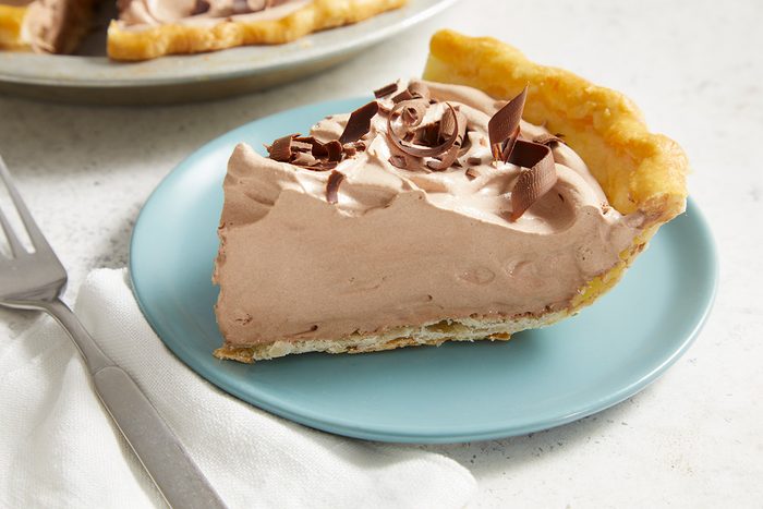 A slice of chocolate cream pie with a flaky crust and chocolate shavings on top, served on a light blue plate next to a fork and a white napkin.