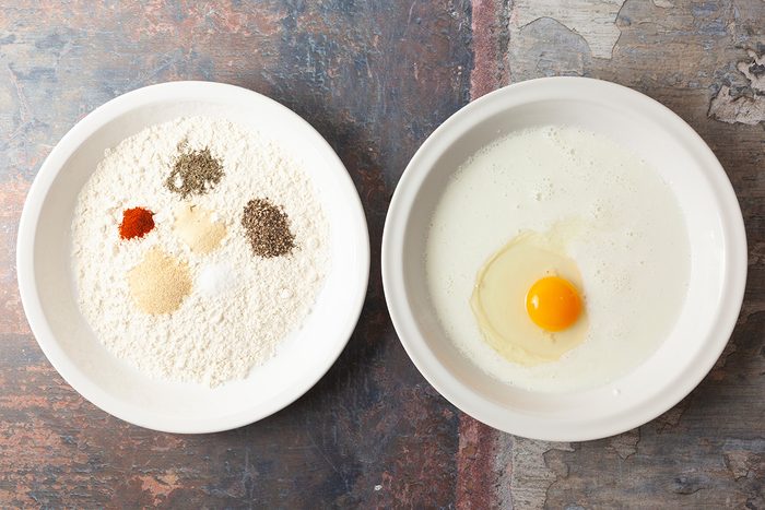 Two white bowls on a textured surface; one bowl contains flour, spices, and seasonings, while the other contains milk and a raw egg.