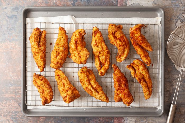 Twelve crispy, golden-brown fried chicken tenders are arranged on a wire rack set over a baking sheet, with a slotted metal spoon nearby. The background surface is lightly textured.