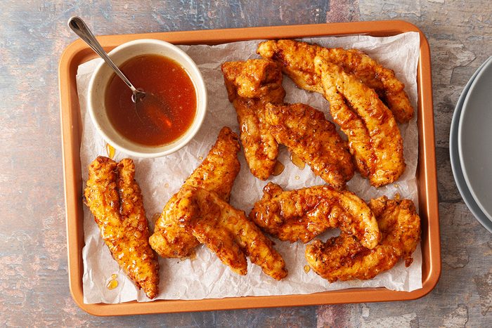 A tray of crispy fried chicken tenders on parchment paper, served with a small bowl of orange-colored dipping sauce and a spoon.
