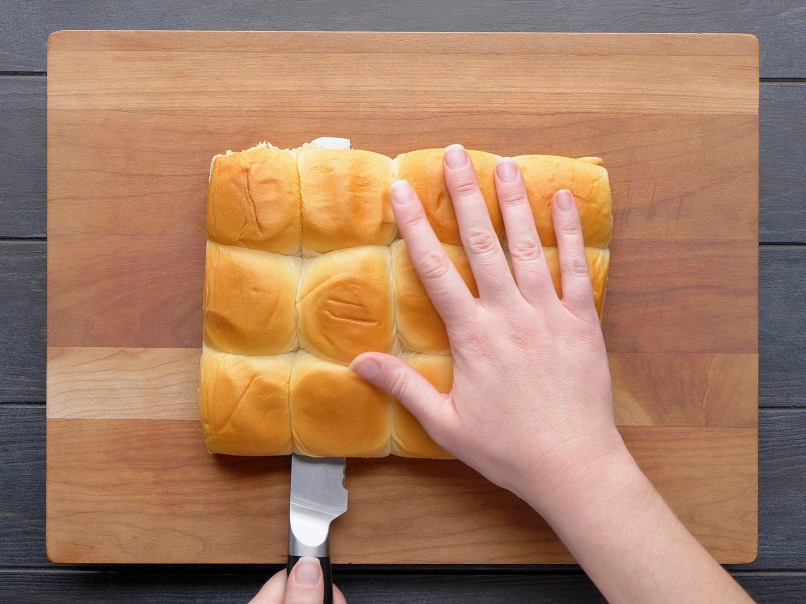A person’s hand holds a block of connected dinner rolls on a wooden cutting board while the other hand slices them horizontally with a bread knife
