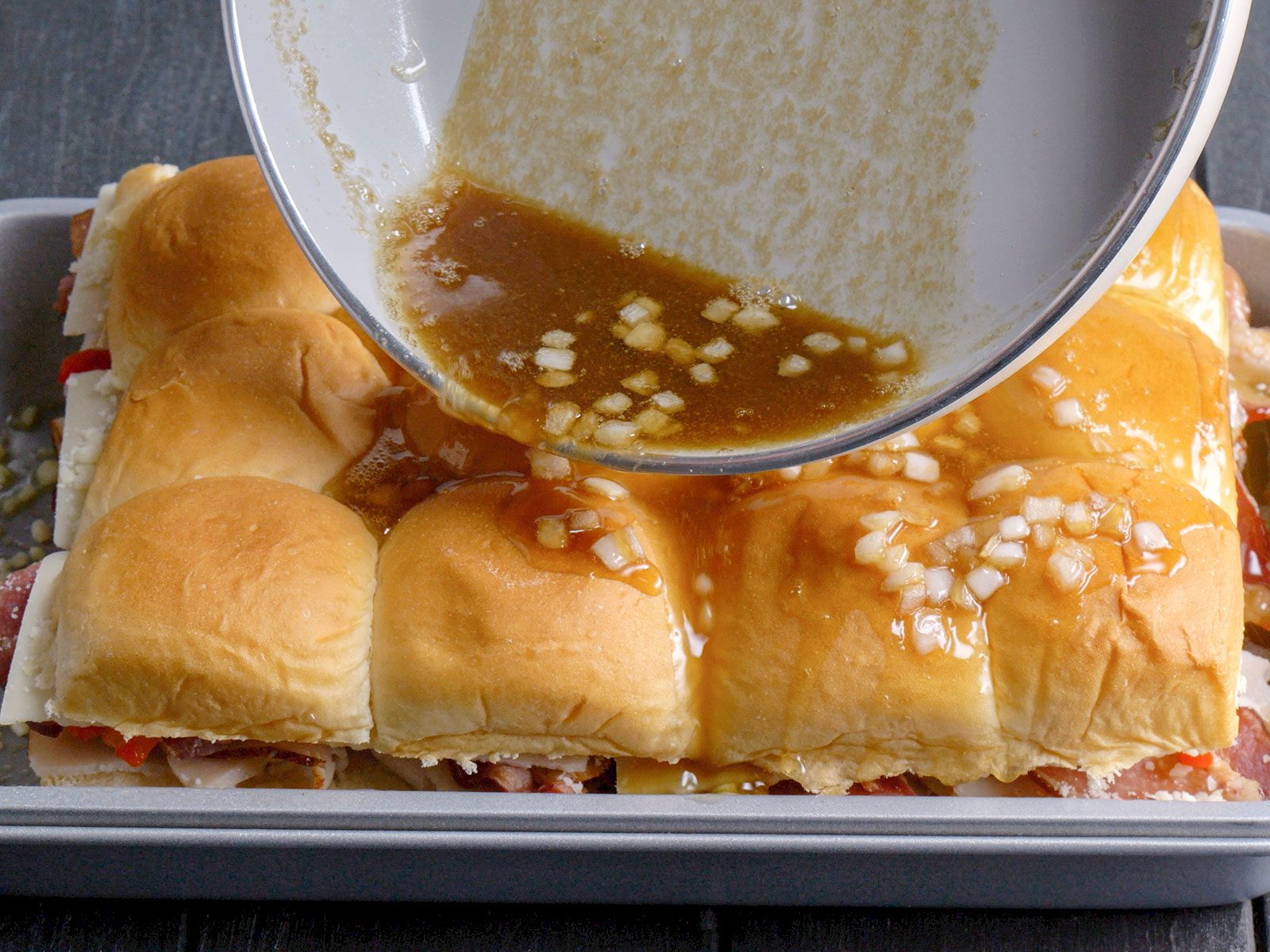 A close-up of a baking tray with slider sandwiches, as a mixture with chopped onions is being poured over the top from a saucepan