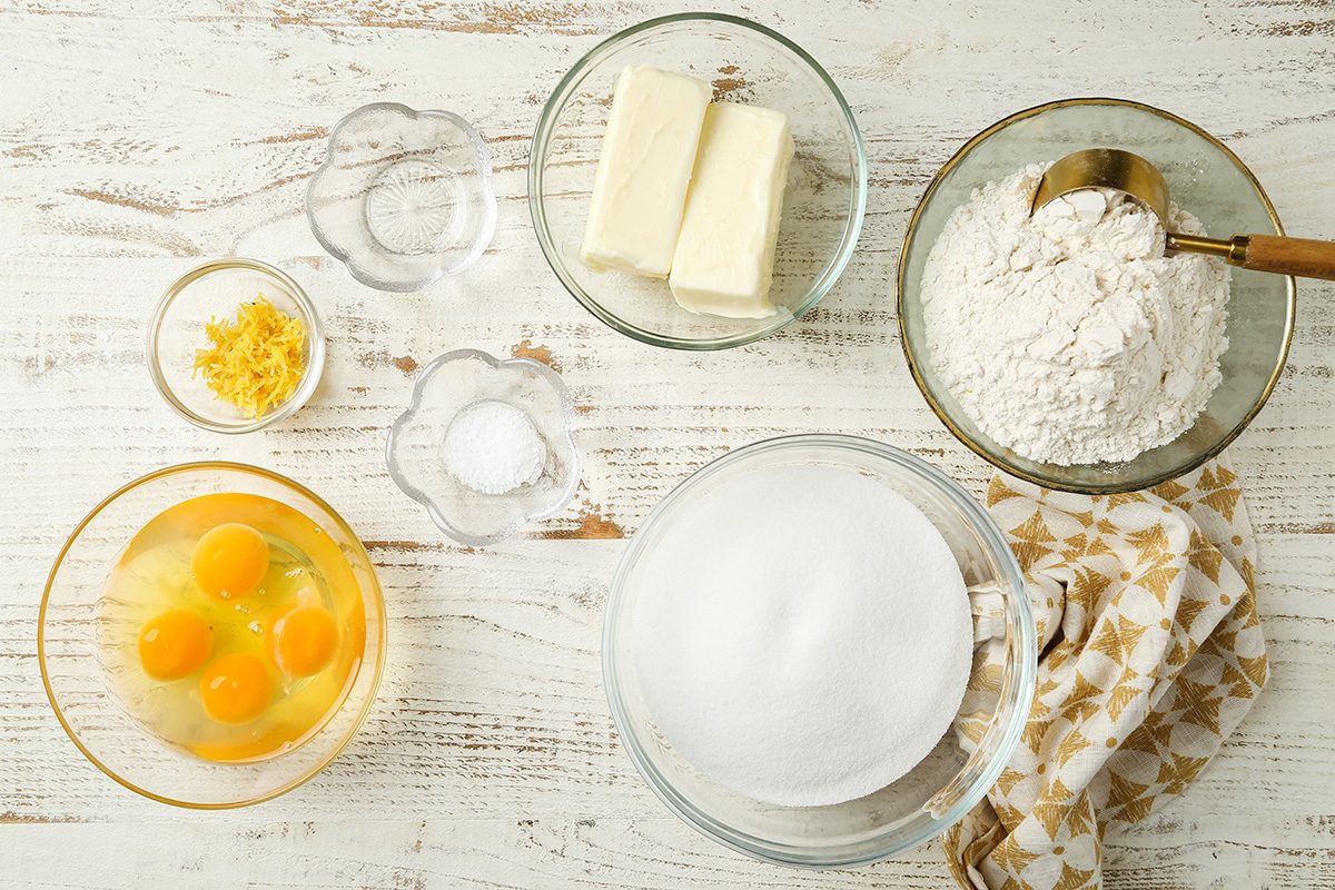 Top view of baking ingredients on a white wooden surface: eggs in a bowl, sticks of butter, flour with a spoon, sugar, salt, baking powder, and lemon zest, with a patterned cloth on the side.
