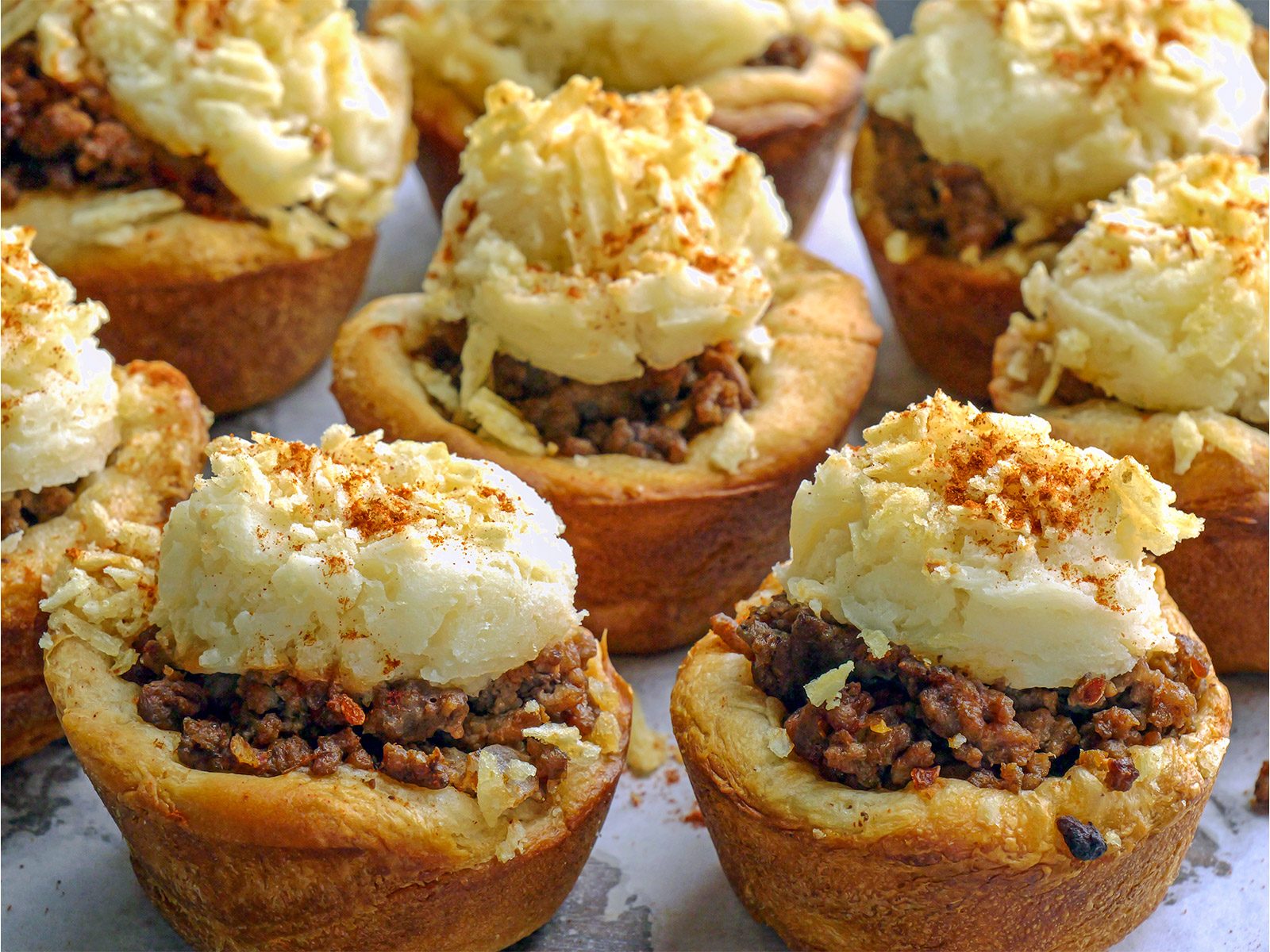 Mini meat pies topped with mashed potatoes, grated cheese, and a sprinkle of seasoning, arranged closely together on a surface. The pies have a golden, flaky crust and a hearty meat filling.