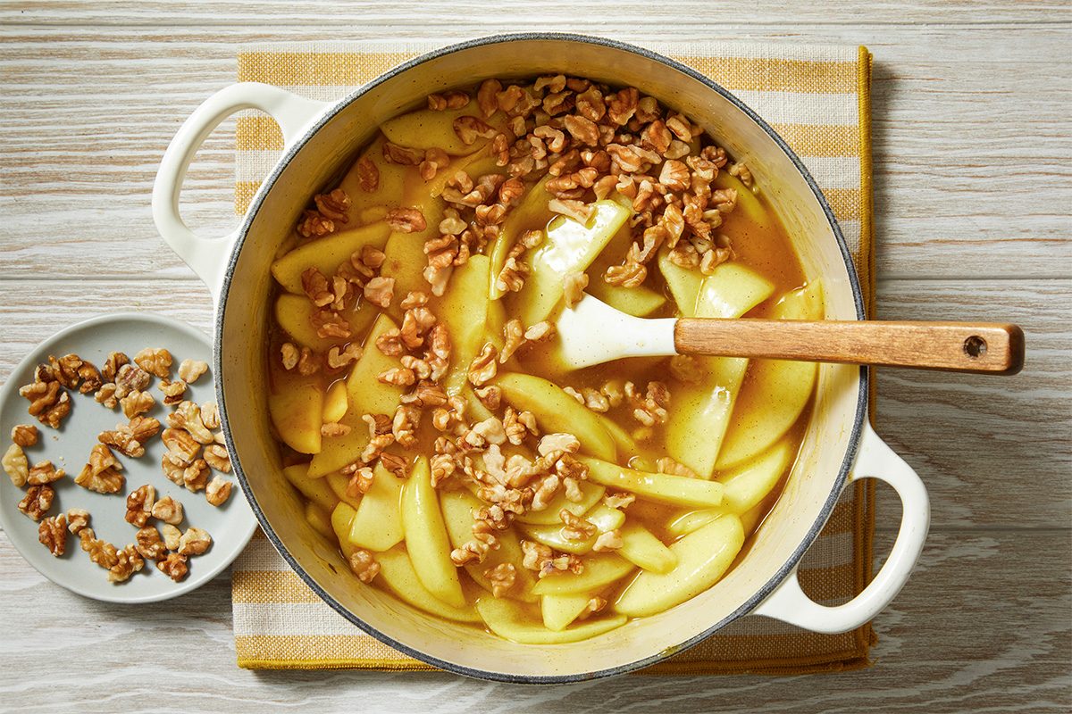 A pot filled with cooked apple slices and walnuts sits on a striped cloth, with a spatula resting inside. A small plate of extra walnuts is placed next to the pot on a wooden surface.