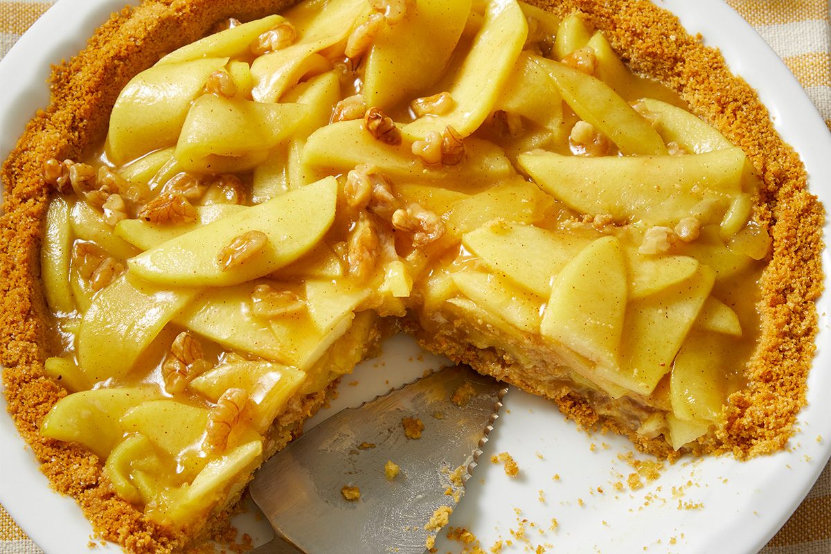 A partially sliced apple pie with a crumbly crust, topped with glossy apple slices and walnuts, sits on a white plate. A serving spatula rests in the empty space where a slice was removed.