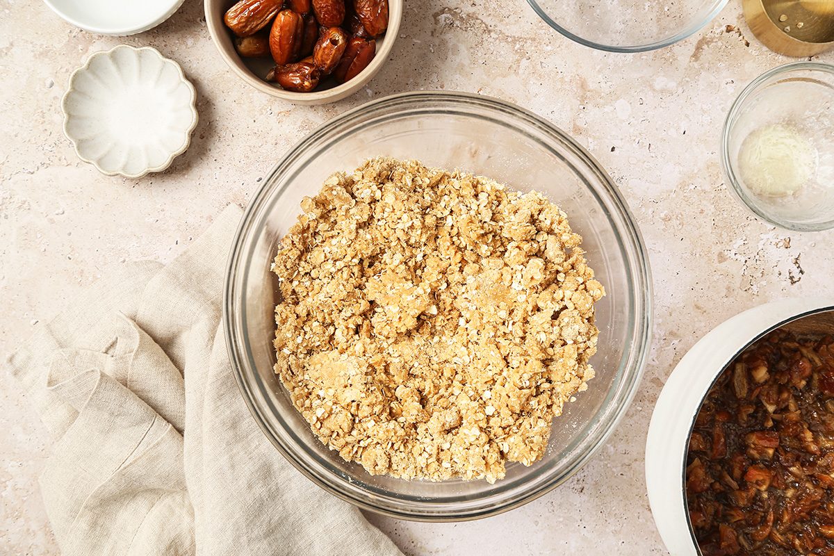 A glass bowl filled with a crumbly oat mixture sits on a light countertop, surrounded by small bowls containing dates, a white powder, and other baking ingredients. A beige cloth is placed nearby.