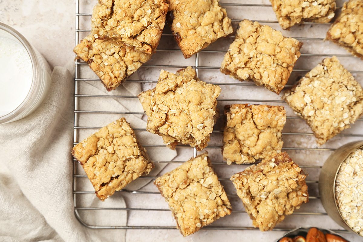 Oatmeal crumble bars are arranged on a cooling rack with a jar of milk nearby. Some loose oats and a bowl of pecans can be seen at the edge of the image on a light background.