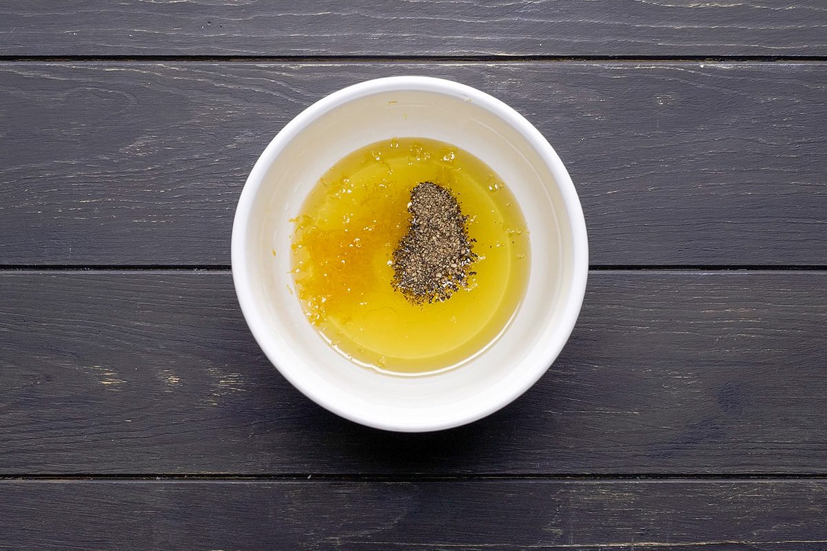A white bowl containing a mixture of yellow liquid and a small mound of ground black pepper sits on a dark wooden surface.
