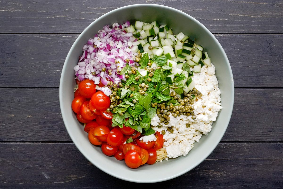 A bowl with chopped red onions, diced cucumbers, halved cherry tomatoes, crumbled cheese, capers, and fresh herbs, arranged in sections on a dark wooden table.