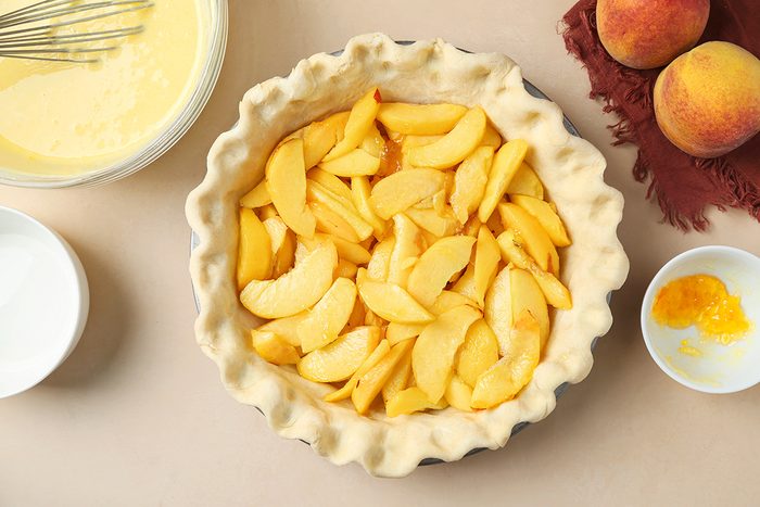Unbaked pie crust filled with sliced peaches on a beige surface, surrounded by a bowl of batter, whole peaches on a cloth, and small bowls of ingredients.