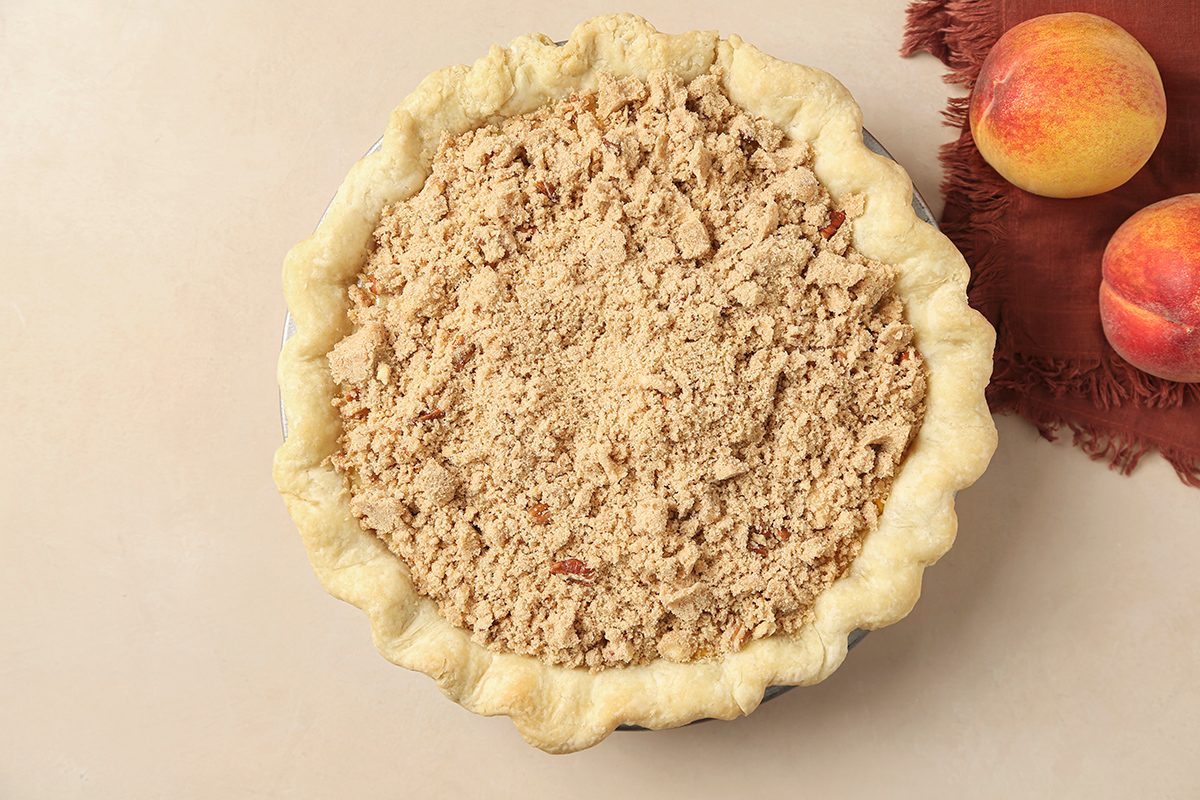 A peach crumb pie with a golden, crimped crust and a crumbly topping sits on a light surface next to two fresh peaches and a brown cloth.