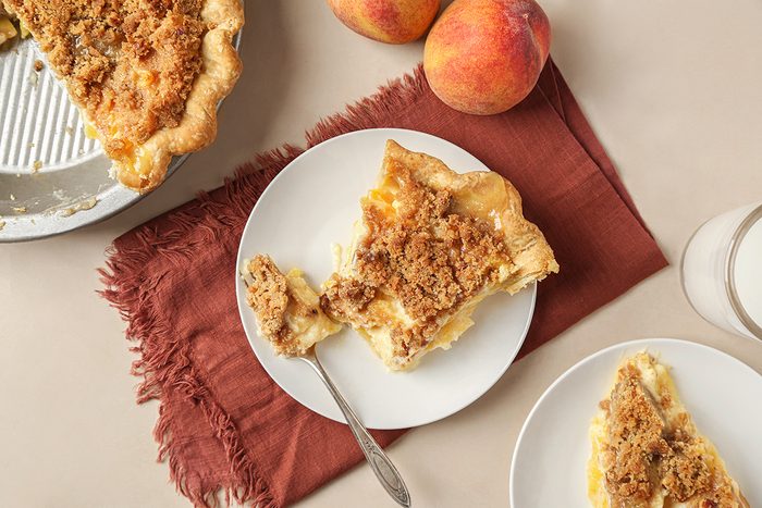 A slice of peach crumble pie on a white plate with a fork, next to a glass of milk, whole peaches, and a pie dish on a beige surface with a rust-colored napkin.