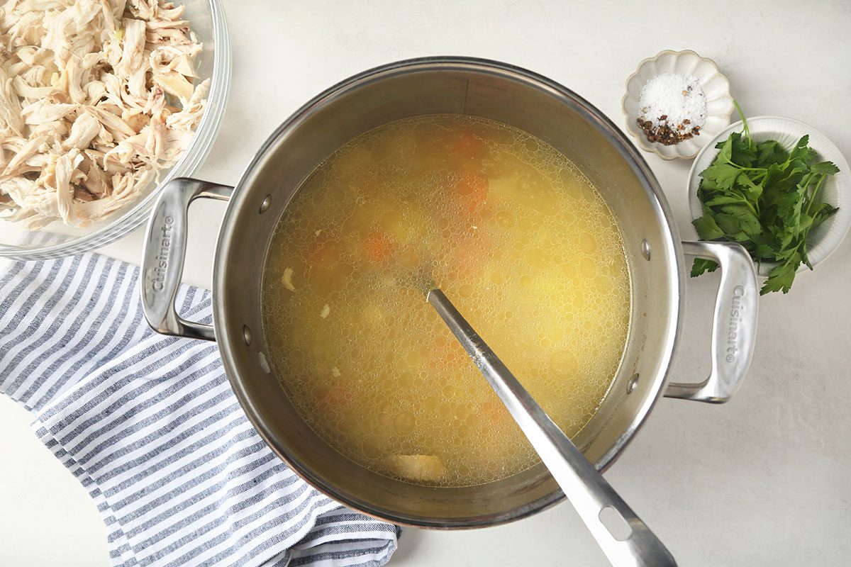A pot of soup with a ladle inside sits on a white surface, surrounded by a bowl of shredded chicken, a dish of leafy greens, a small plate with peppercorns, and a striped kitchen towel.