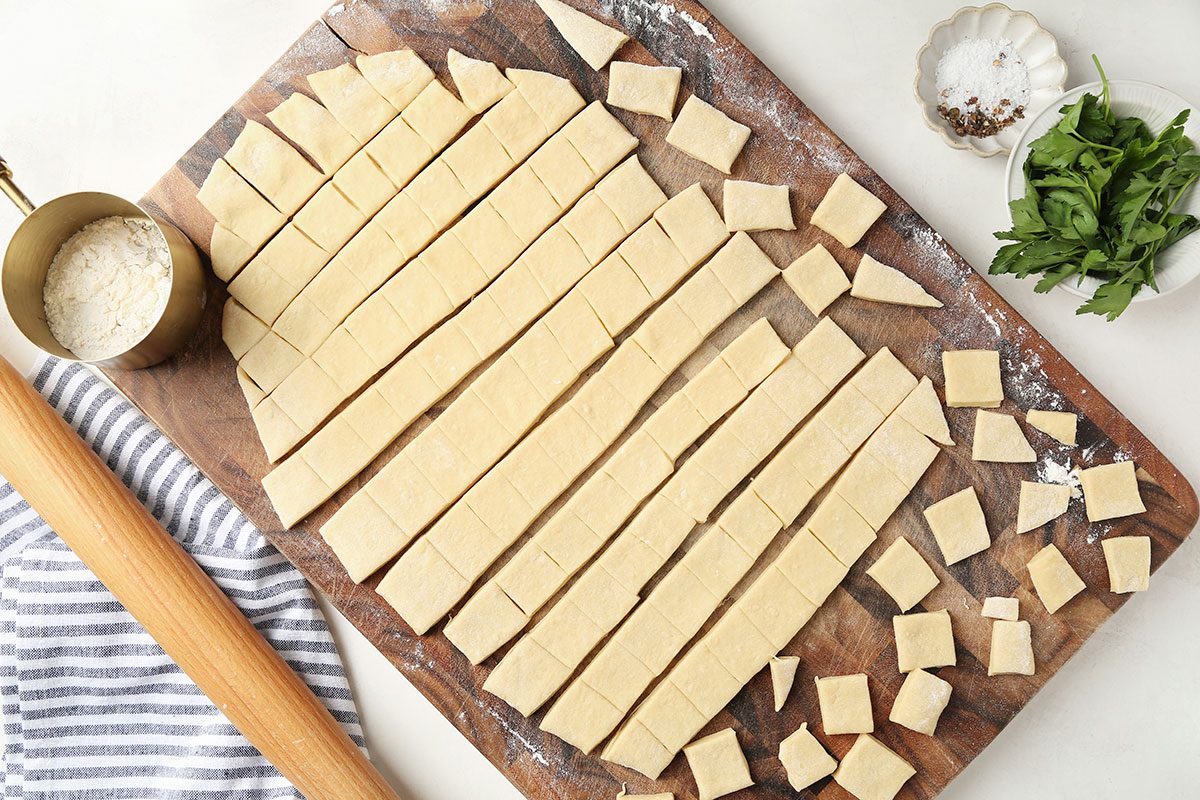 A wooden board with strips and squares of uncooked dough, a rolling pin, a cup of flour, a bowl of salt, and a pile of fresh greens on a white surface.