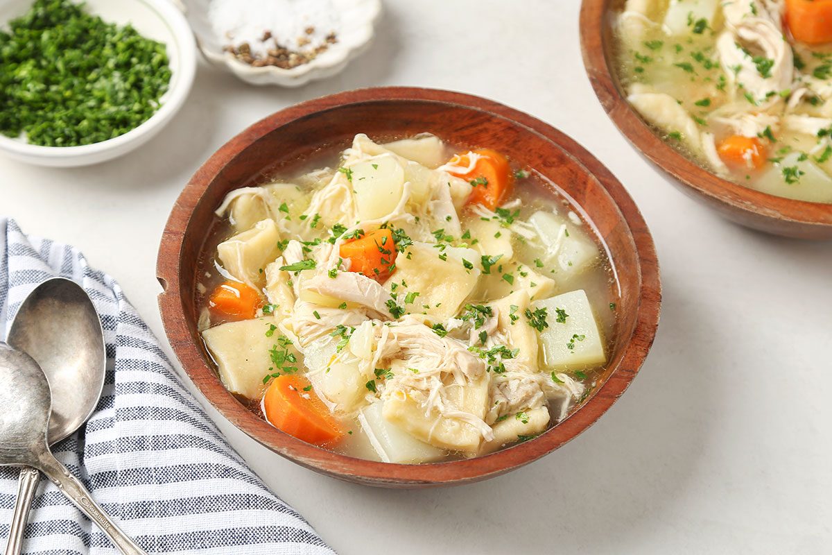 A wooden bowl filled with chicken soup containing chunks of chicken, potatoes, carrots, and celery, garnished with fresh herbs. A side bowl of chopped herbs and a striped napkin with spoons are nearby.