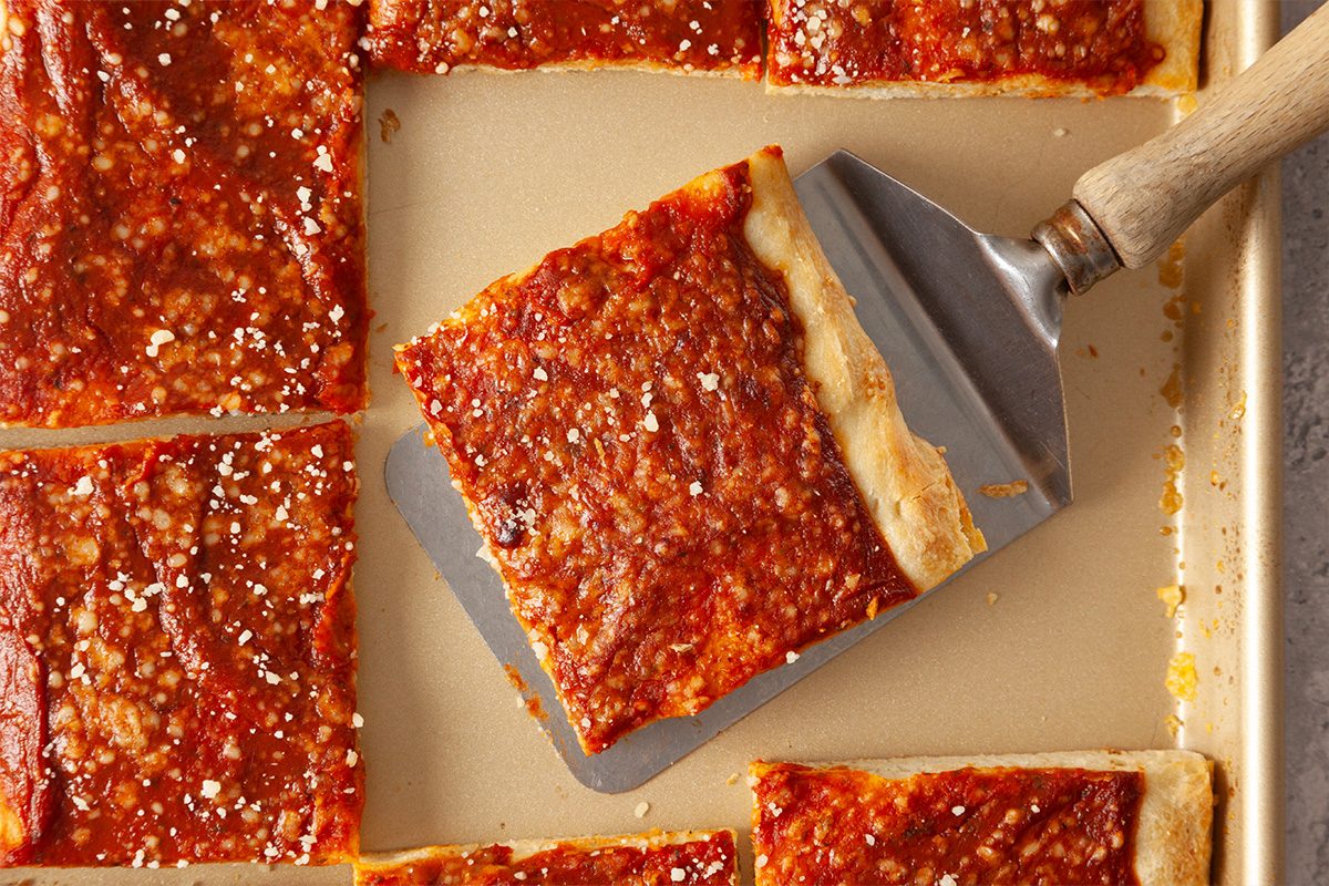 A metal spatula lifts a square slice of pizza with a thick, golden-brown crust and tomato sauce, surrounded by other pizza slices on a baking sheet.