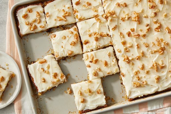 A baking tray with frosted sheet cake cut into squares, topped with chopped walnuts. Some pieces are separated, and a plate holds one square piece. The cake has a creamy white frosting and a light brown base.
